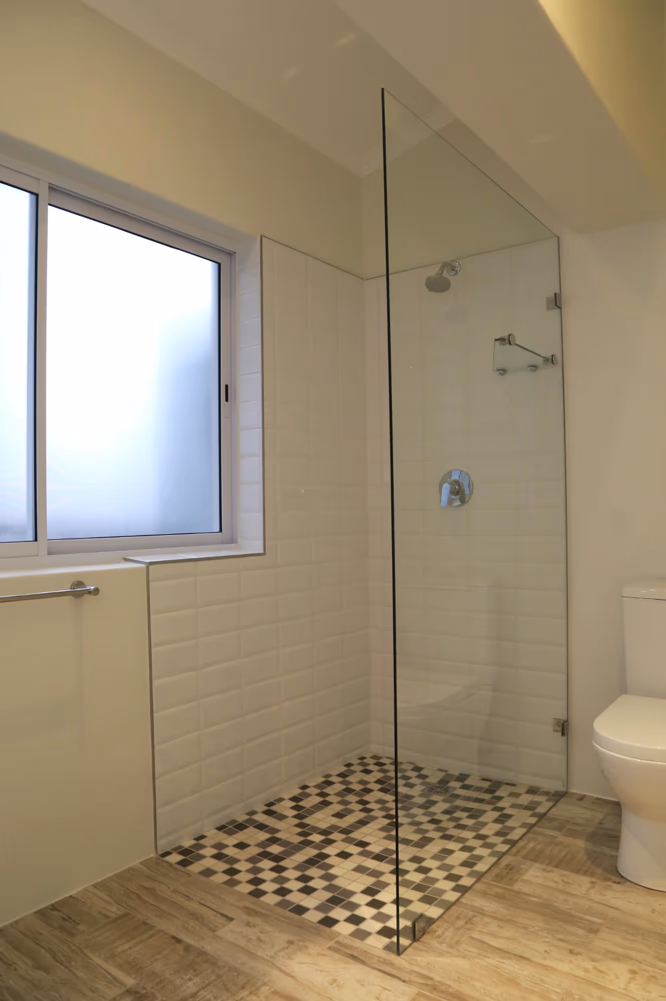 Bathroom with a glass walk-in shower featuring checkered black and white floor tiles, white subway tiles on the walls, a frosted window, and a white toilet. Work by Wricon construction.