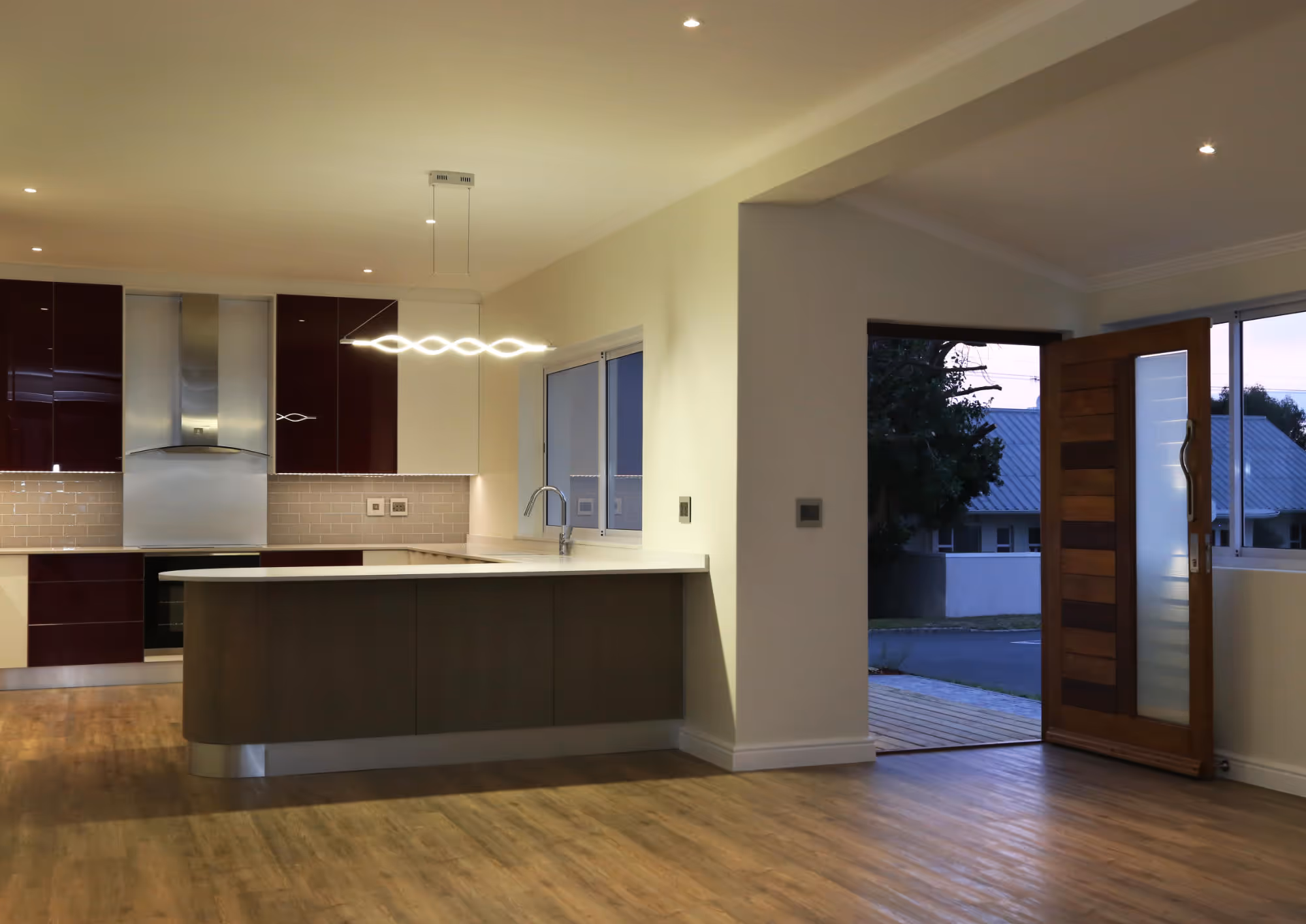 Modern kitchen with wooden floor, maroon and white cabinets, illuminated wave-shaped pendant light, and an open wooden front door showing a street view at dusk. Work by Wricon construction.