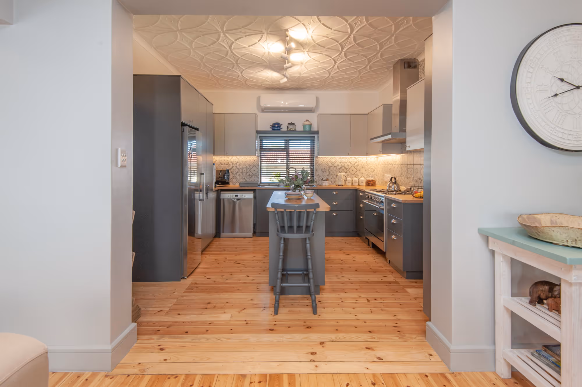 Modern kitchen with grey cabinets, patterned tile backsplash, wooden floor, central island with a chair, and stainless steel appliances. Work by Wricon construction.