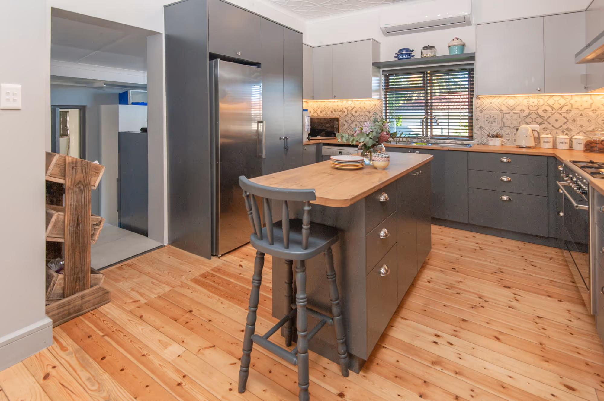 Modern kitchen with gray cabinets, wooden floors, an island with a tall chair, and a window with blinds above the sink. Work by Wricon construction.
