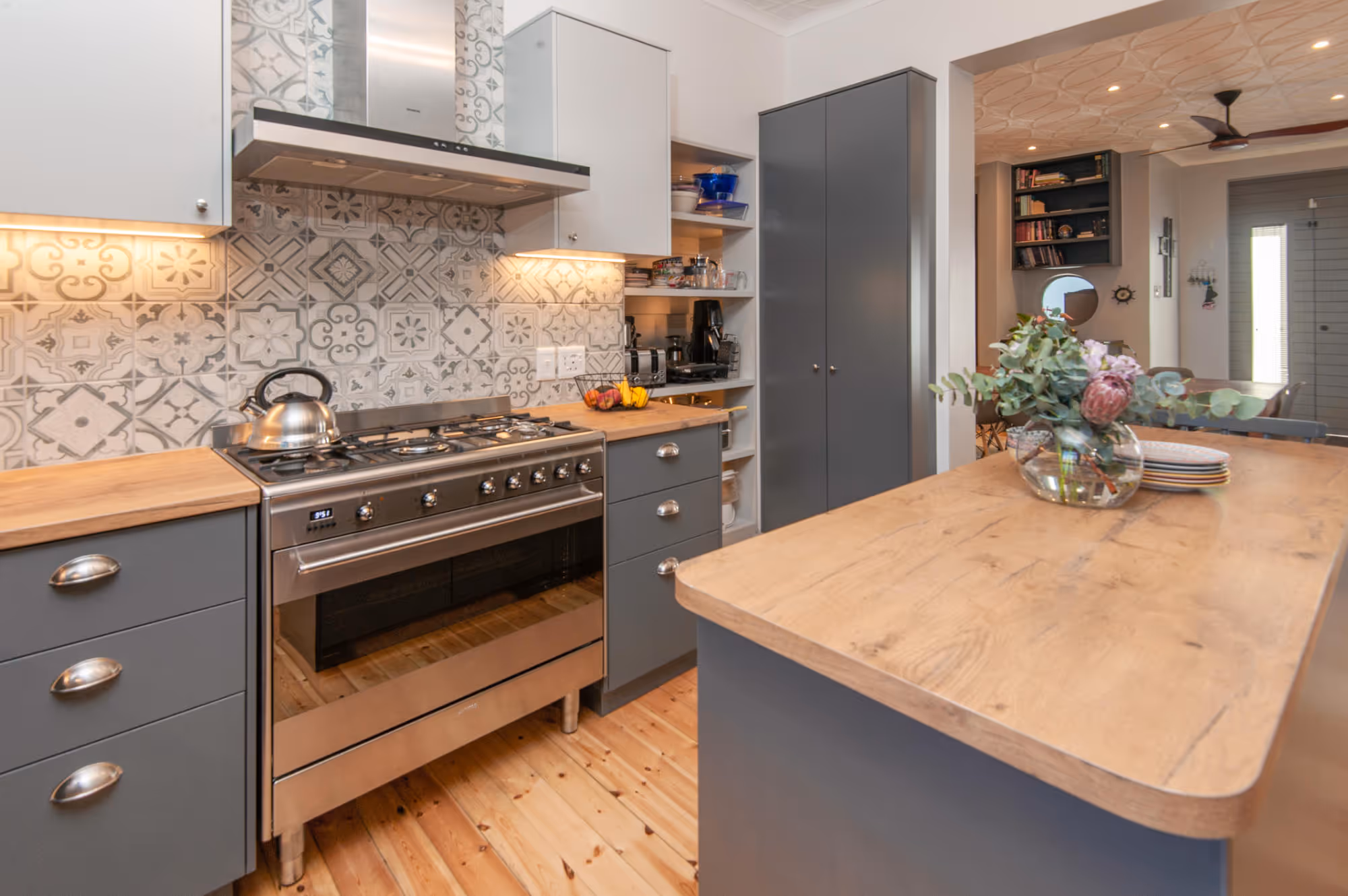 Modern kitchen with gray cabinets, wood countertops, patterned tile backsplash, stainless steel stove with kettle, and a wooden island with a floral vase. Work by Wricon construction.