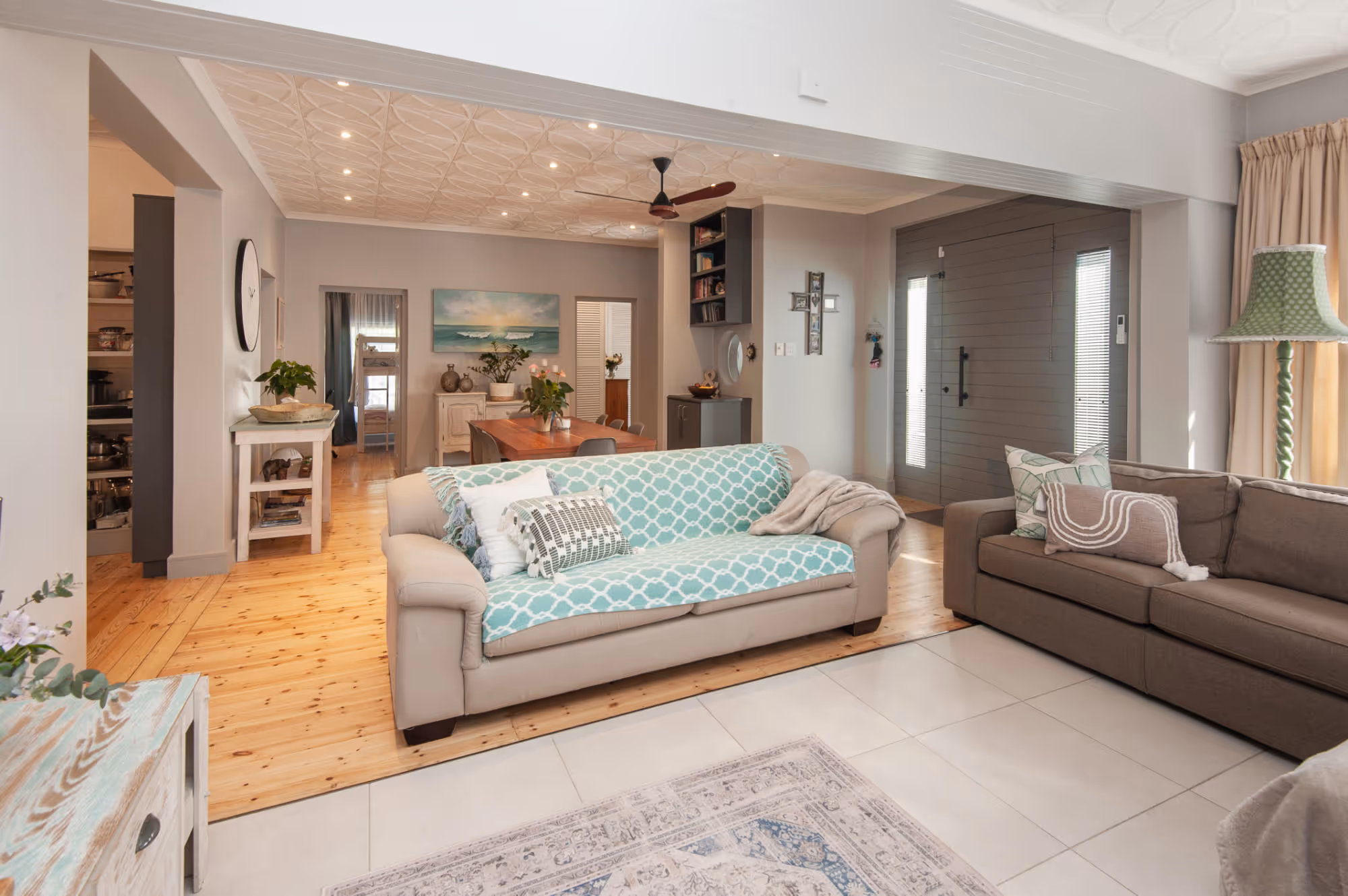Cozy living room with two sofas, one beige with a blue patterned cover and one brown, wooden floor leading to a dining area with a wooden table and chairs, ceiling fan, and a gray front door with sidelights. Work by Wricon construction.