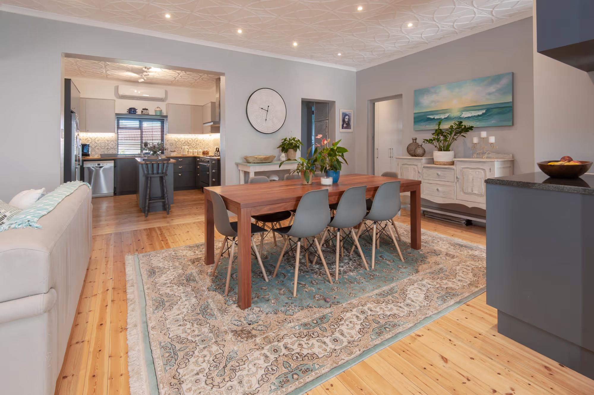 Modern dining area with a wooden table and six gray chairs on a patterned rug, next to a living room and an open kitchen.