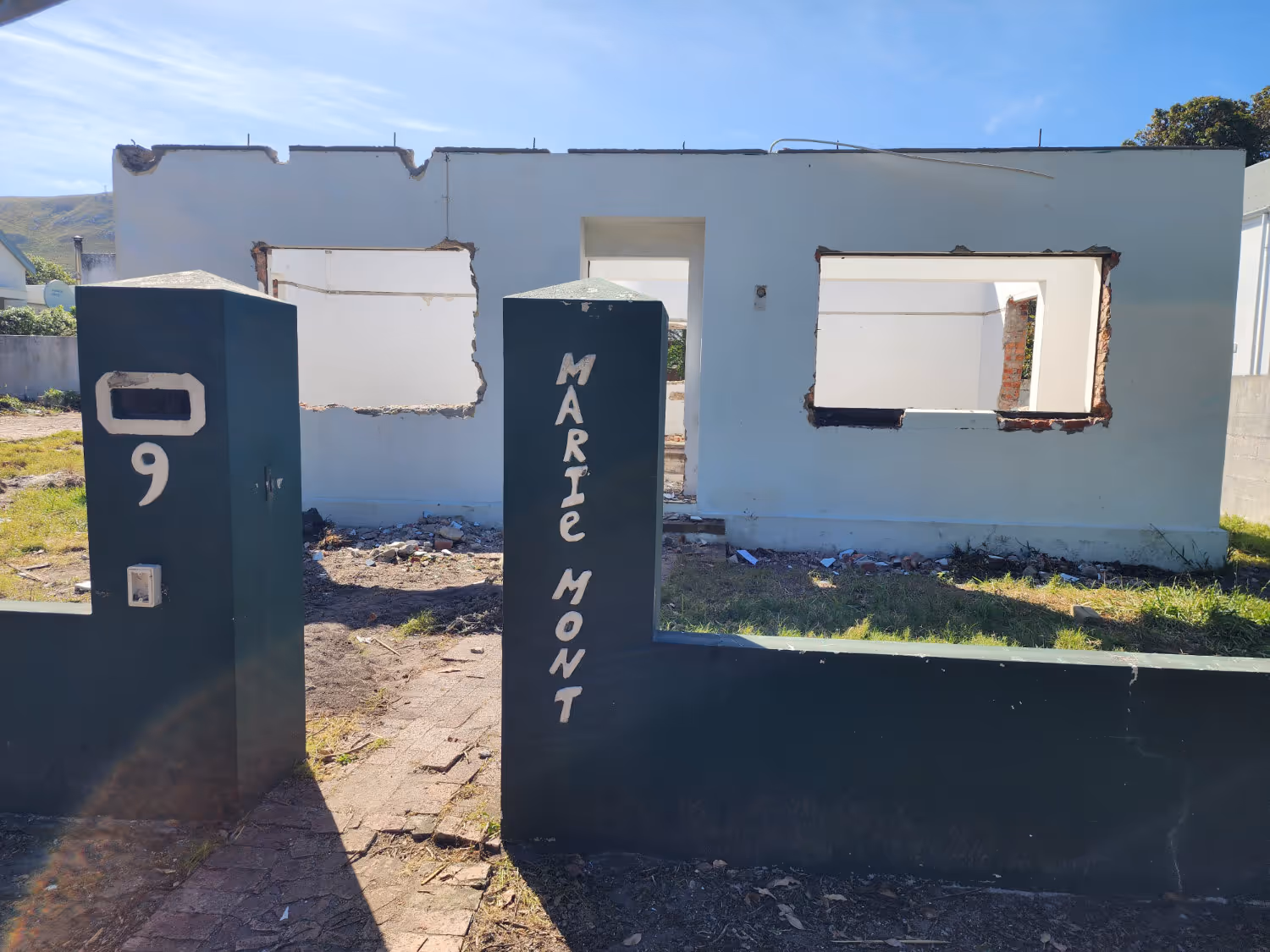 Partially demolished white house visible through a dark green broken perimeter wall with 'Marie Mont' and the number 9 painted on its pillars.