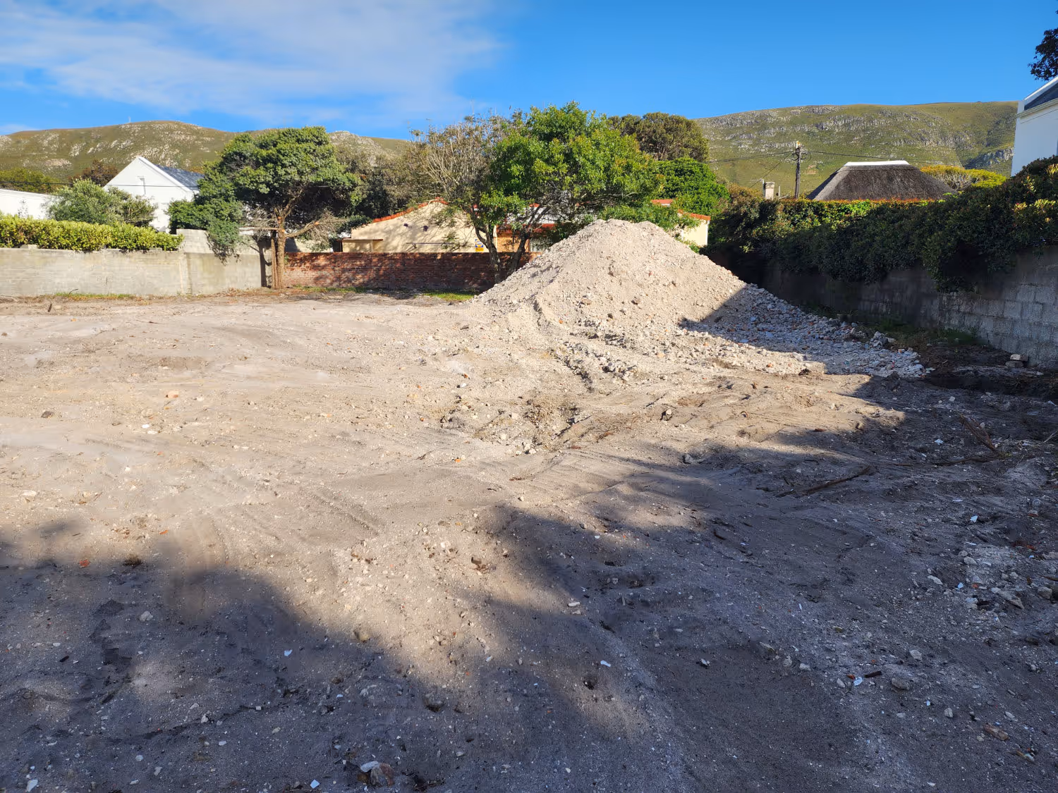 Cleared dirt lot with a large pile of rubble in the middle, surrounded by trees and houses under a blue sky with distant hills.