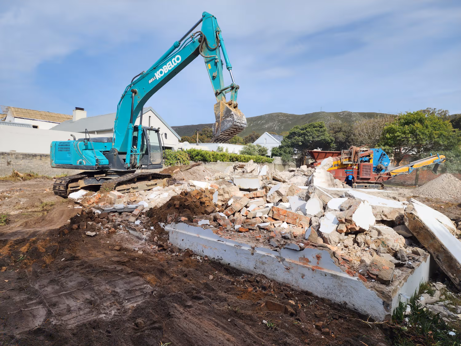 Excavator demolishing a house with rubble and construction debris on the ground under a clear sky.