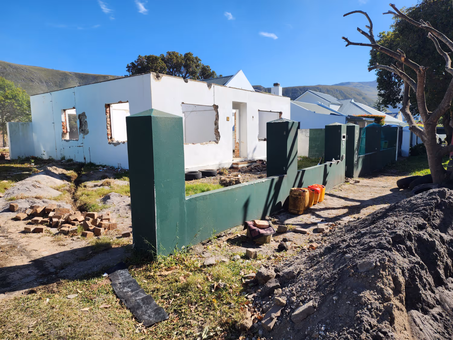 Partially demolished white house with missing windows and bricks scattered in front, bordered by a green fence under a clear blue sky.