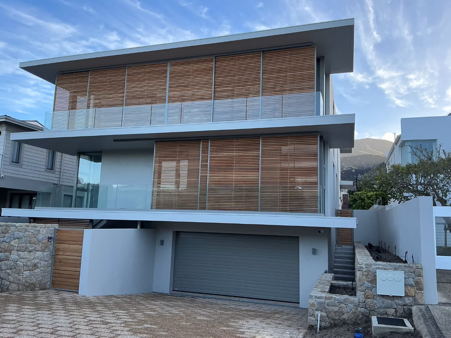 Modern three-story house with wooden slats, glass balconies, and stone fence under a partly cloudy sky.