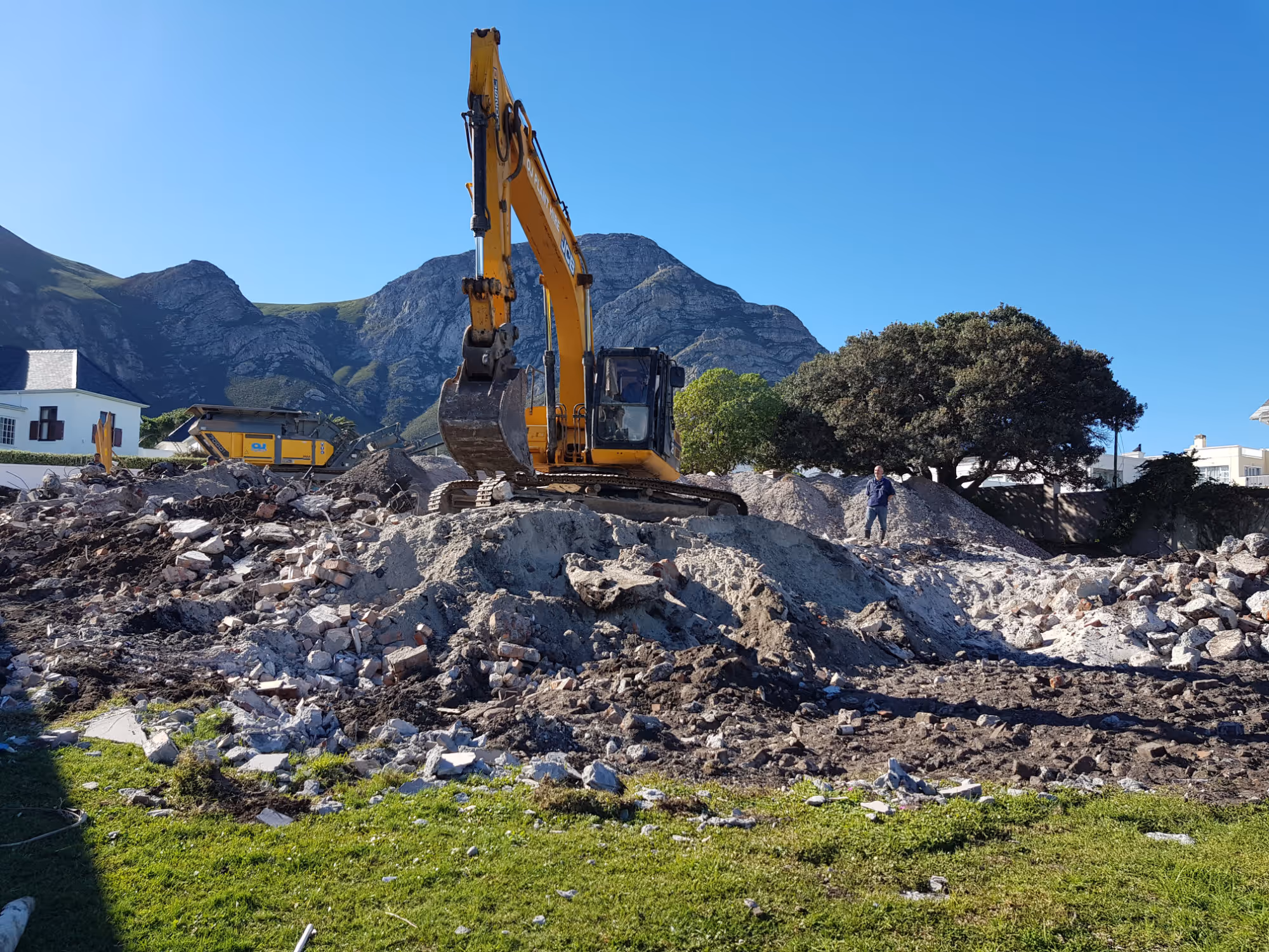 Yellow excavator working on a construction site with rubble and debris, mountains and houses in the background under a clear blue sky. Project by Wricon Construction.