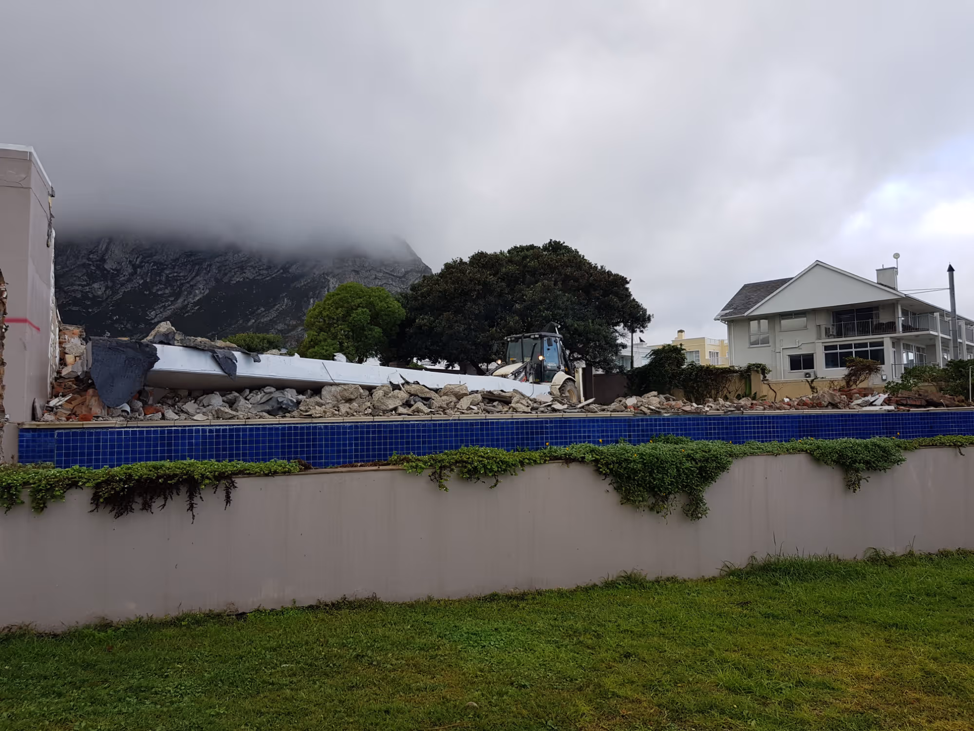 Demolition site with rubble and a backhoe loader near a tiled wall, residential houses, and mountains under cloudy sky. Project by Wricon Construction.