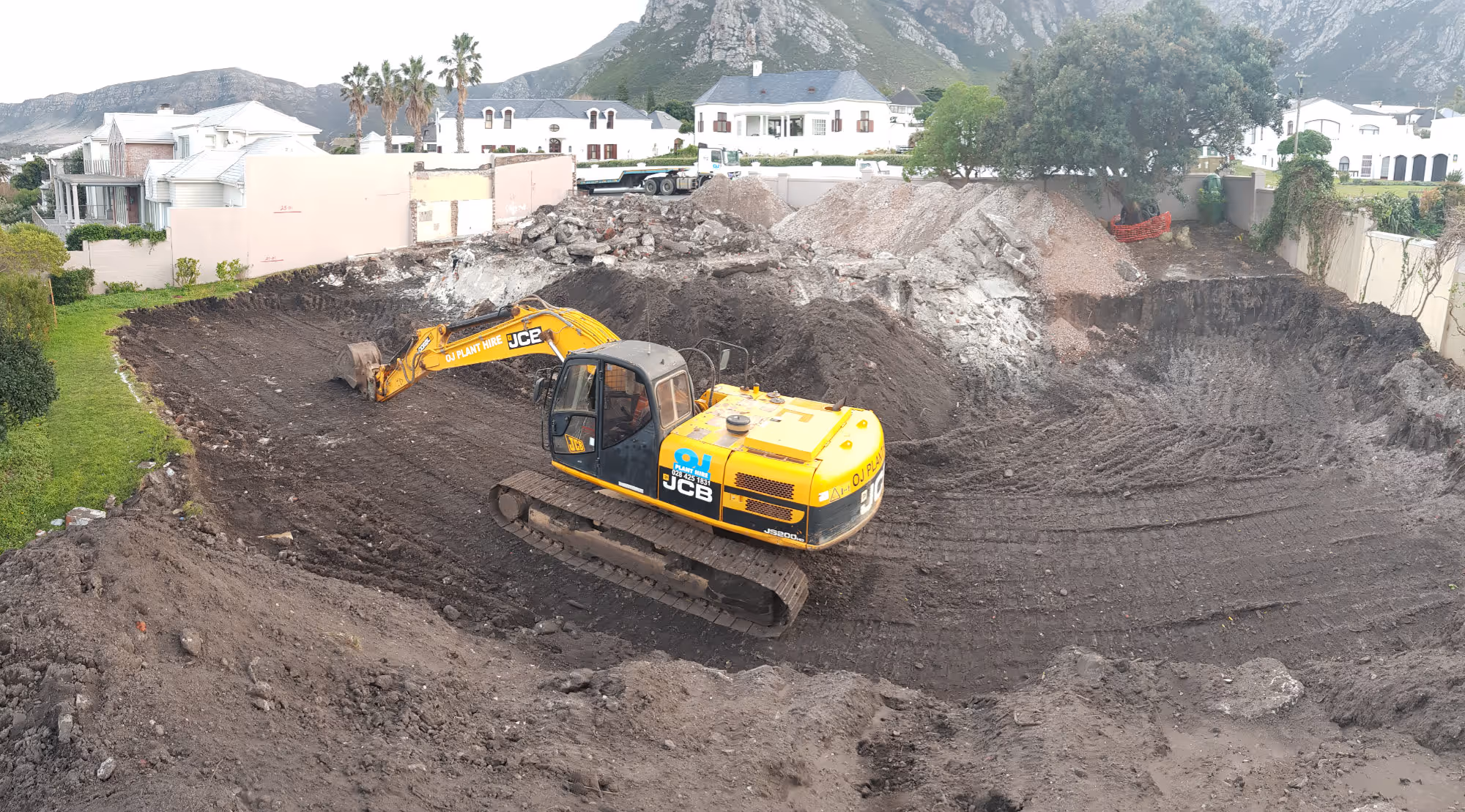Yellow JCB excavator in a large dug-out construction site surrounded by residential houses and mountains. Project by Wricon Construction.