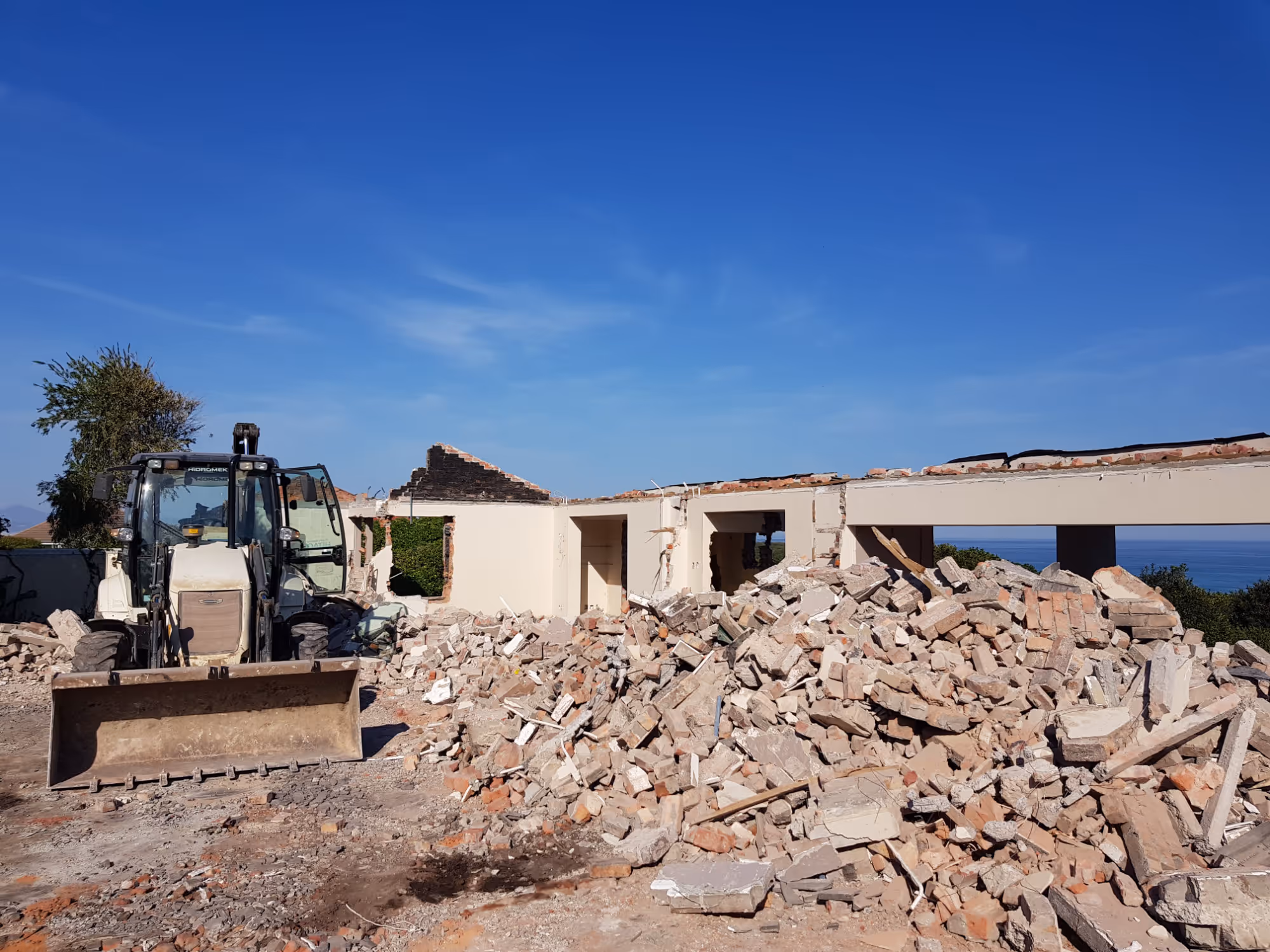 Demolition site with a pile of rubble and a bulldozer in front of a partially demolished building under a clear blue sky. Project by Wricon Construction.