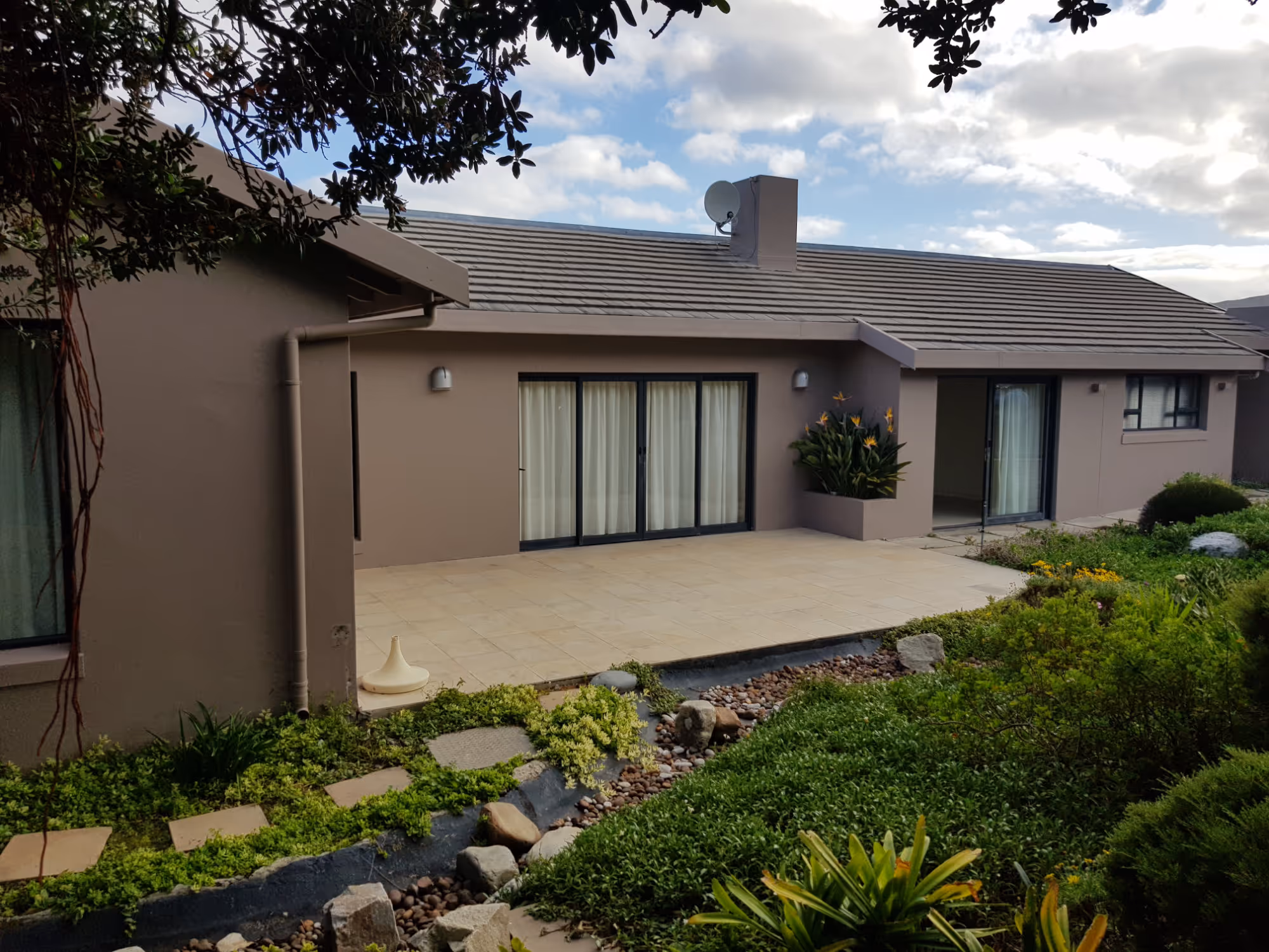 Modern single-story house with beige walls, tiled patio, garden with green shrubs and flowering plants, under a partly cloudy sky. Project by Wricon Construction.