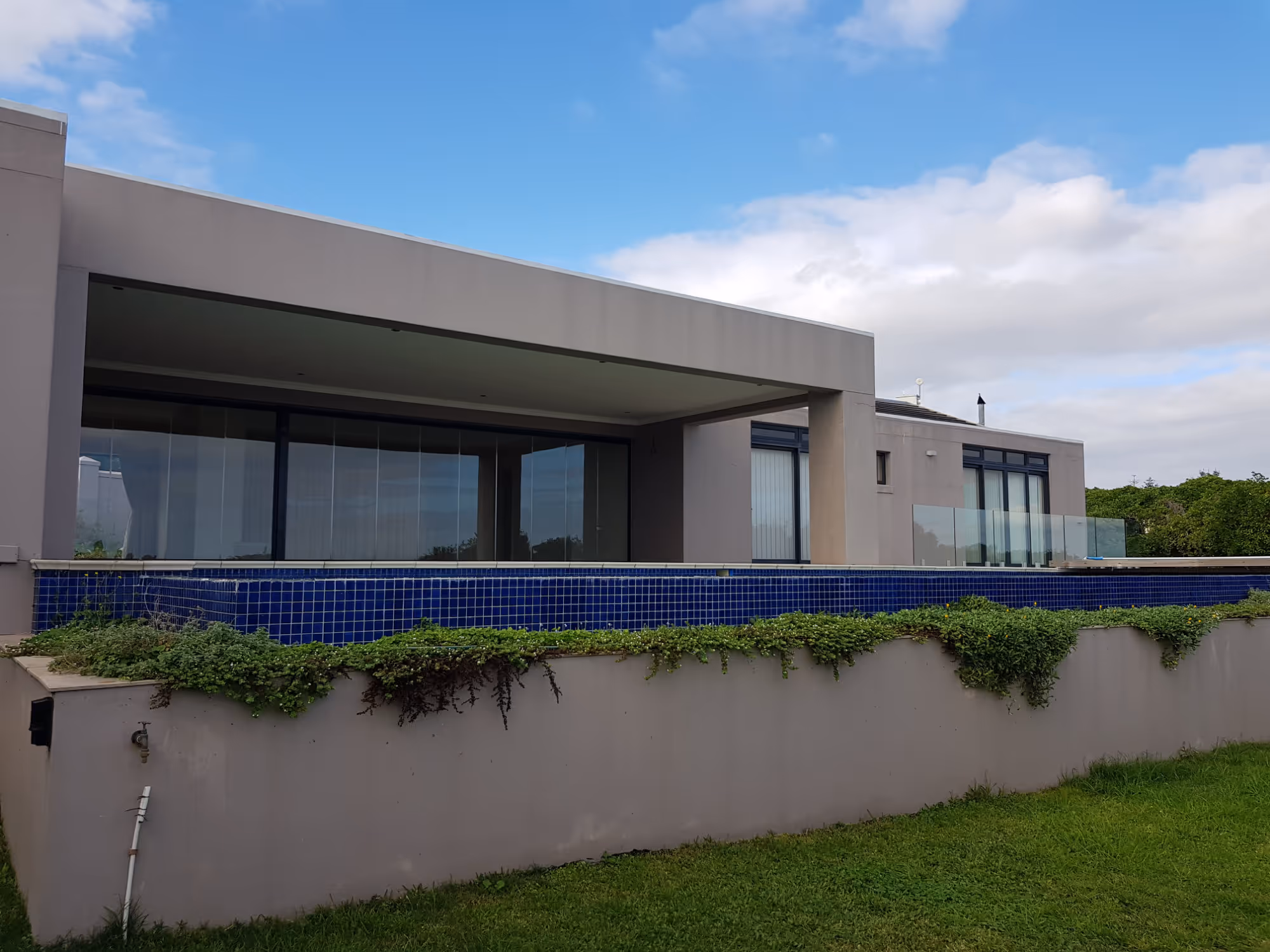 Modern house with large glass windows and a blue-tiled infinity pool surrounded by greenery on a cloudy day. Project by Wricon Construction.