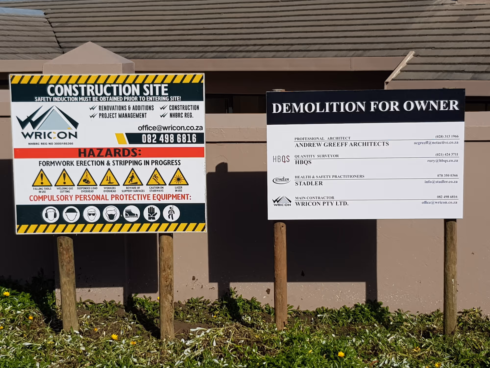 Two construction signs on wooden posts in front of a beige wall, one detailing safety hazards and equipment requirements, the other listing professionals and contractors for a demolition project. Project by Wricon Construction.