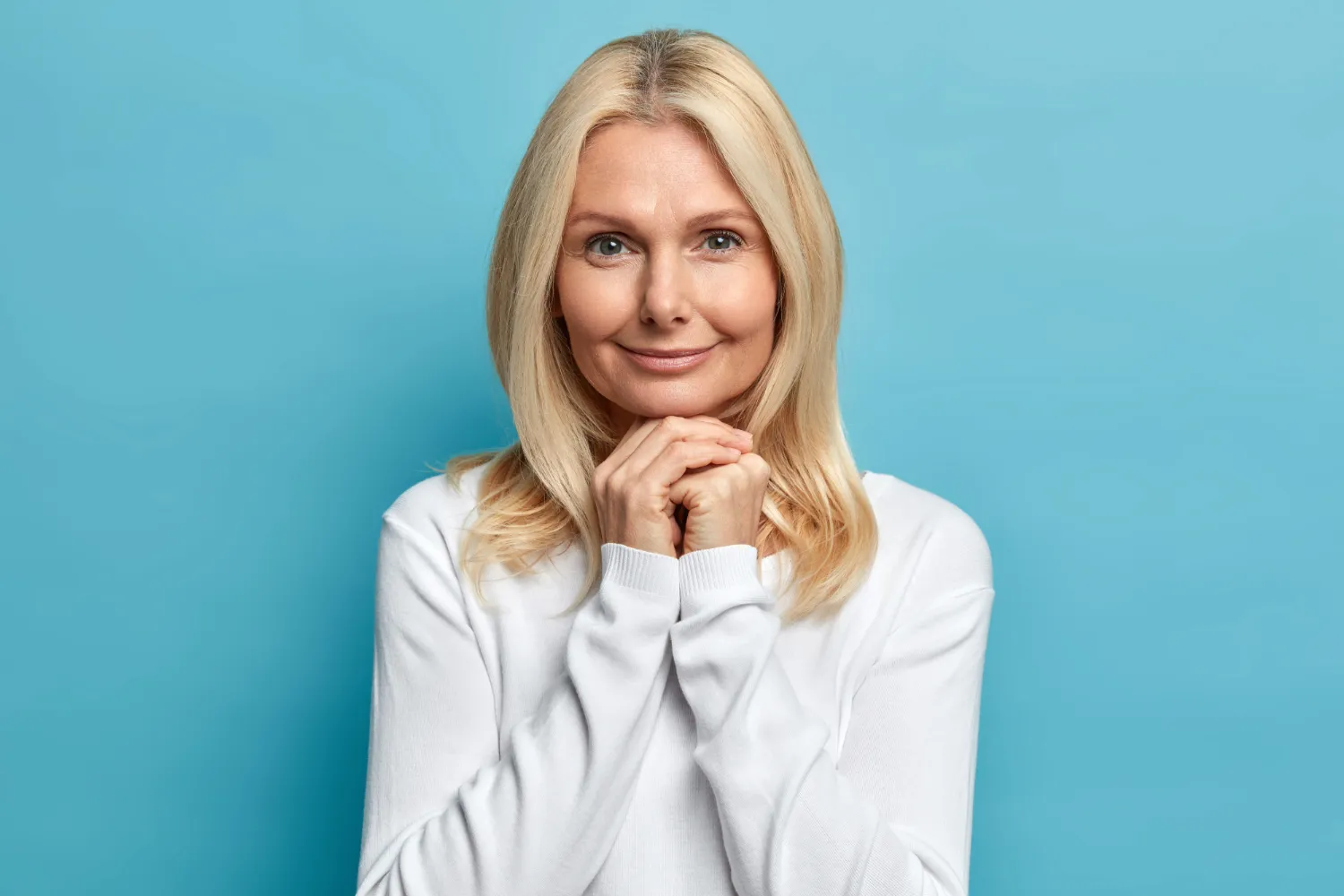 Smiling woman in a white sweater against a blue background, hands clasped under chin, looking serene.