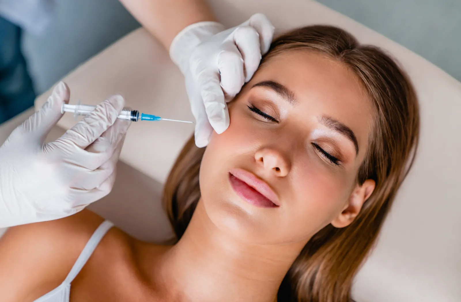 Woman receiving cosmetic injection in cheek, eyes closed, wearing white top, administered by gloved professional.