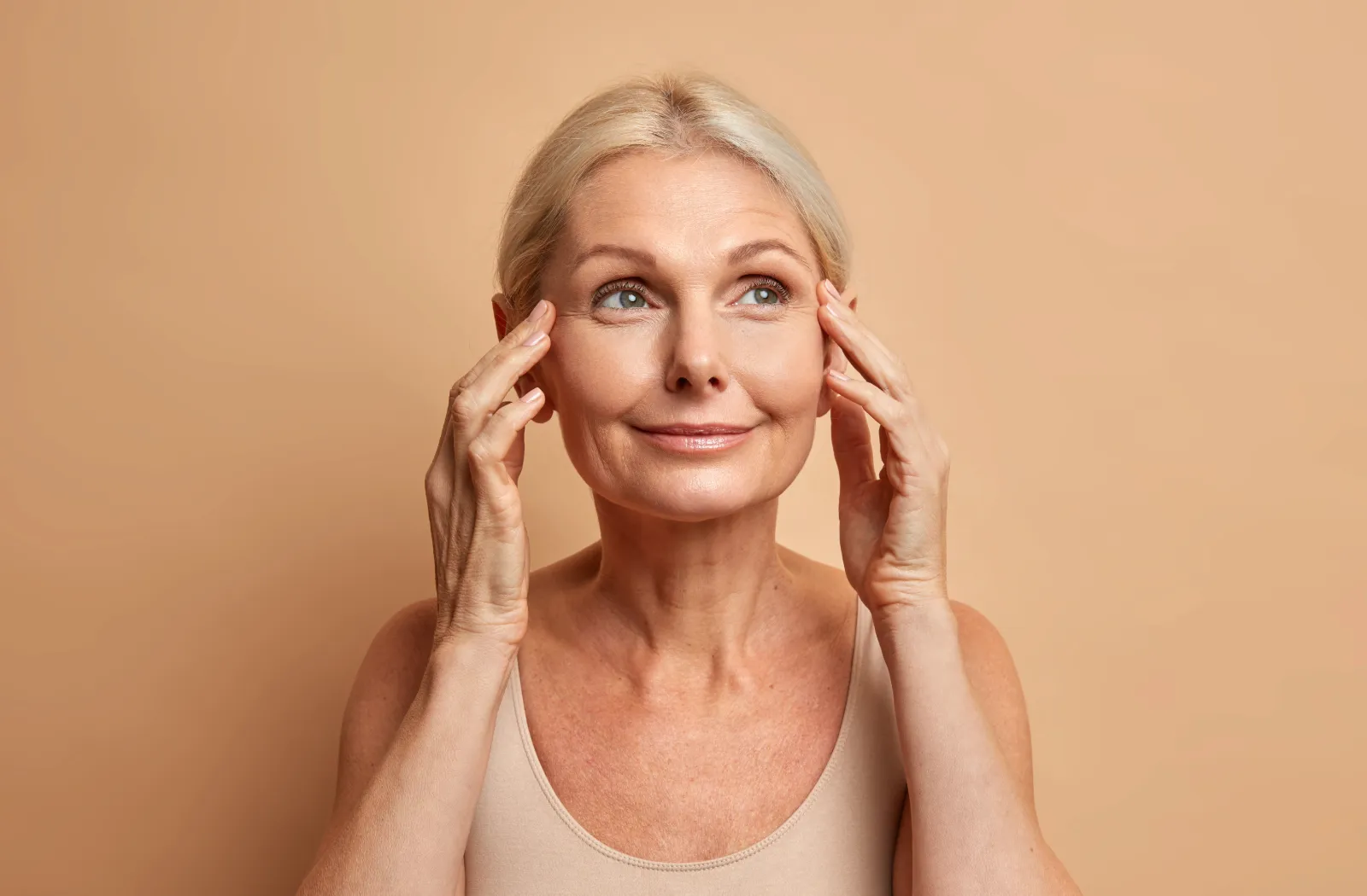 Elderly woman with short blond hair smiling, touching temples, against a beige background.