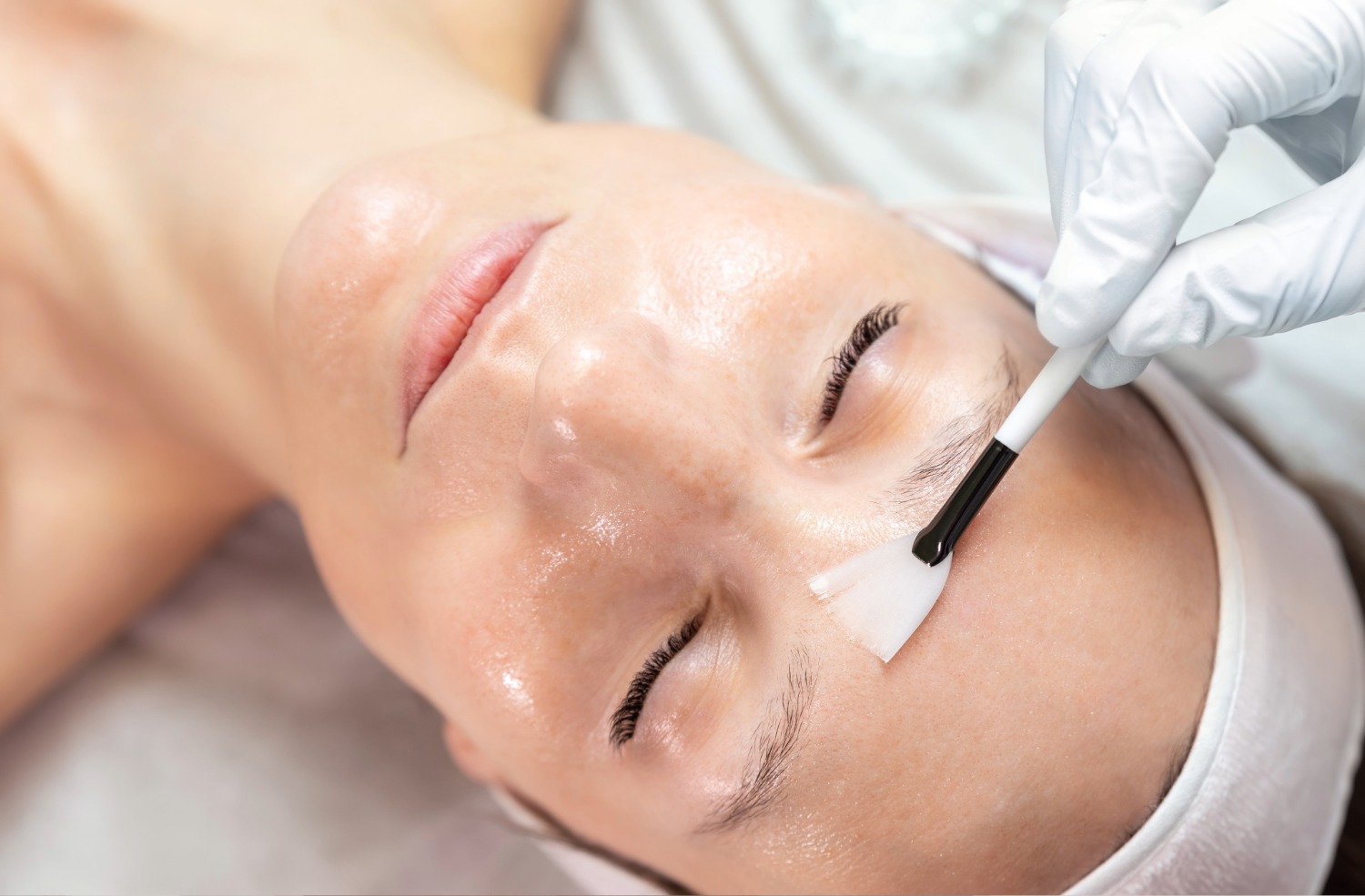 Close-up of a person with closed eyes receiving a facial treatment with a brush applying a product to the forehead.