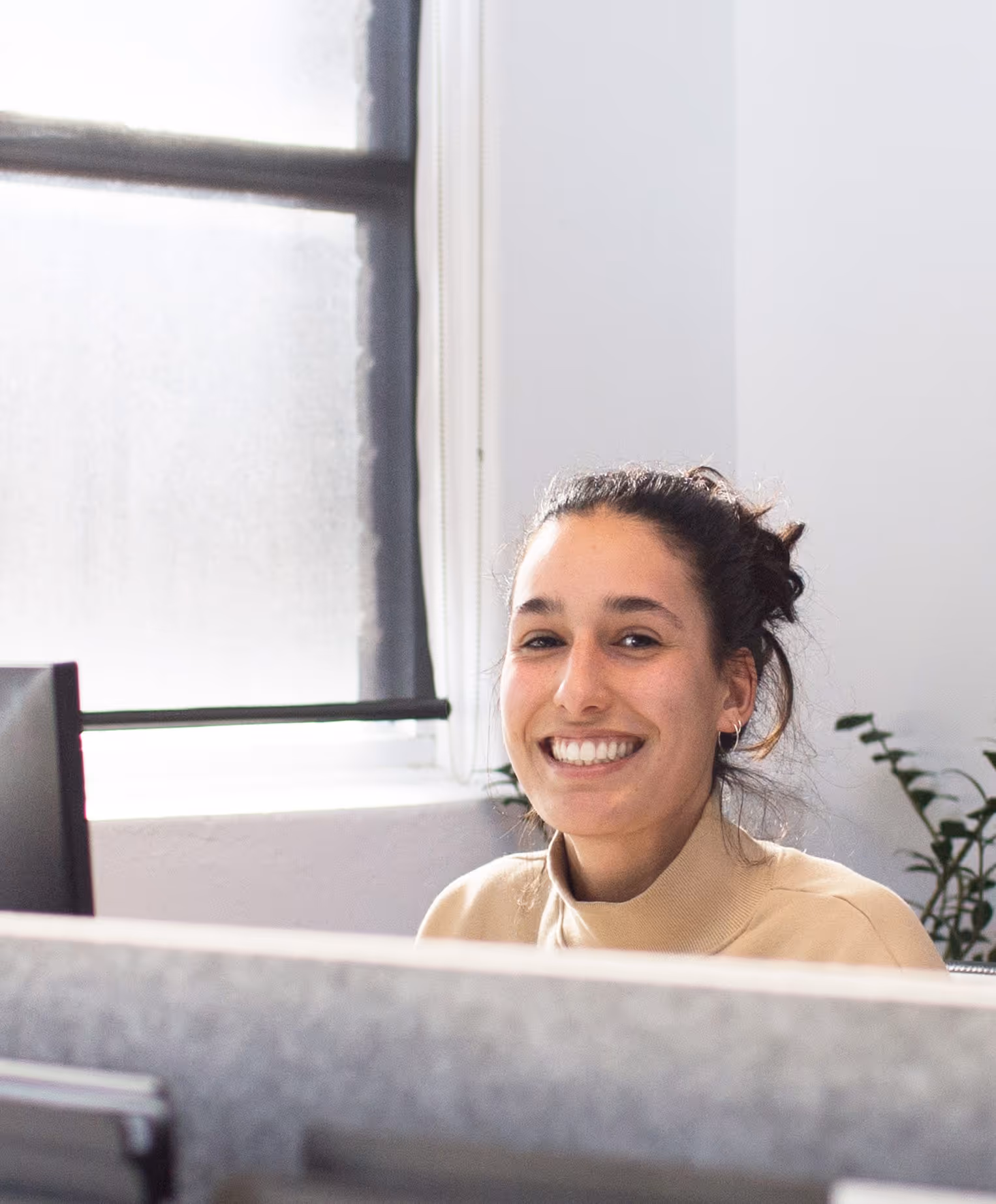 Smiling woman with dark hair in a bun sitting at an office desk with a window and plants in the background.
