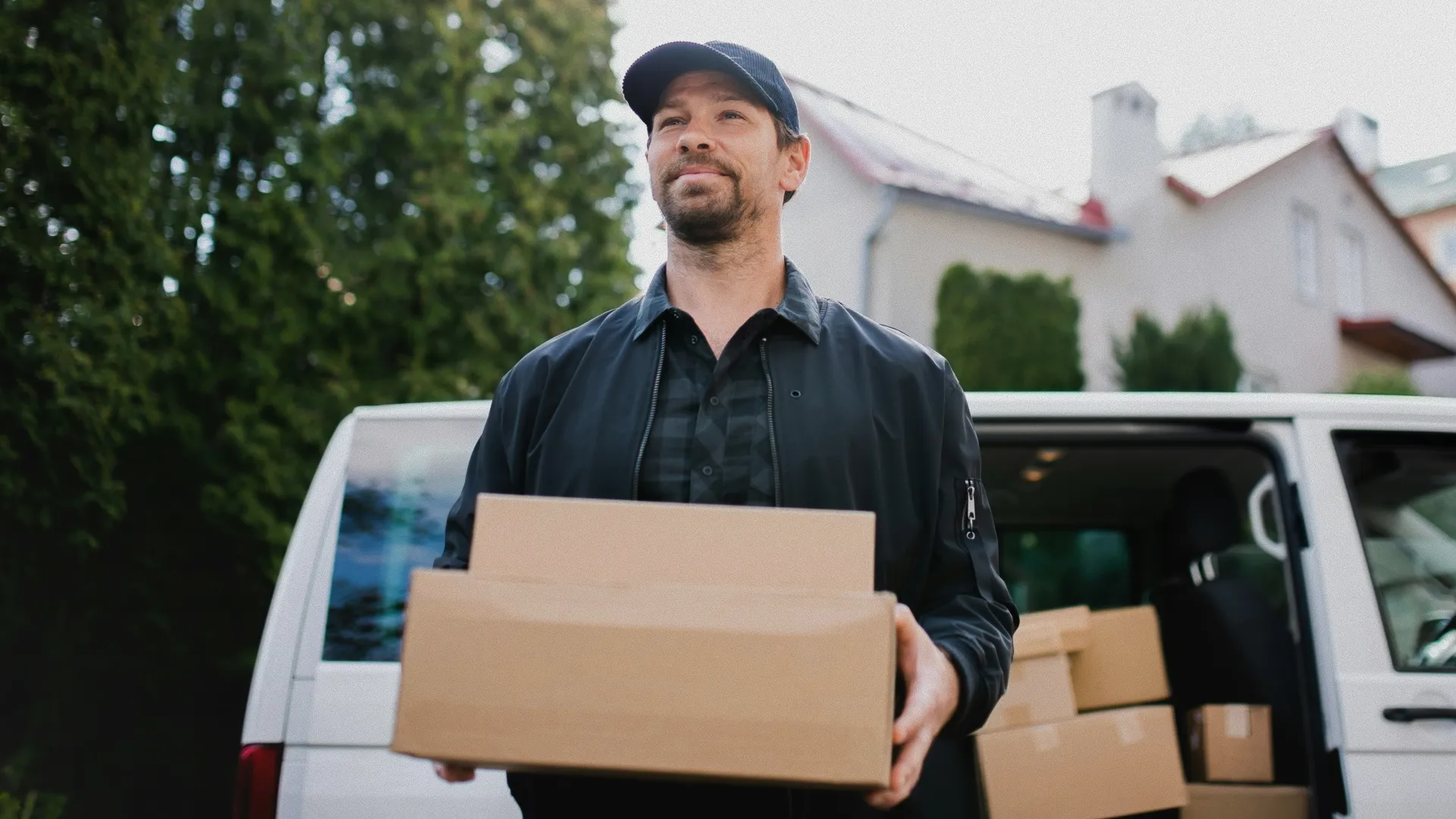 A delivery driver carrying cardboard boxes in front of a white van outside a house