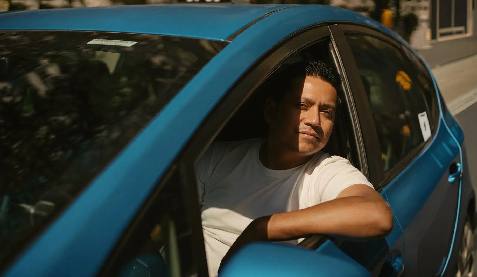 Man with short black hair wearing a white t-shirt, sitting in the driver's seat of a blue car, leaning his arm out the open window and looking ahead with a relaxed expression in sunlight.