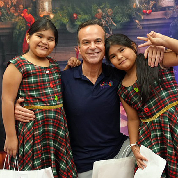 Jorge Plasencia in a navy shirt smiling and posing with two girls in matching red and green plaid dresses holding white shopping bags.