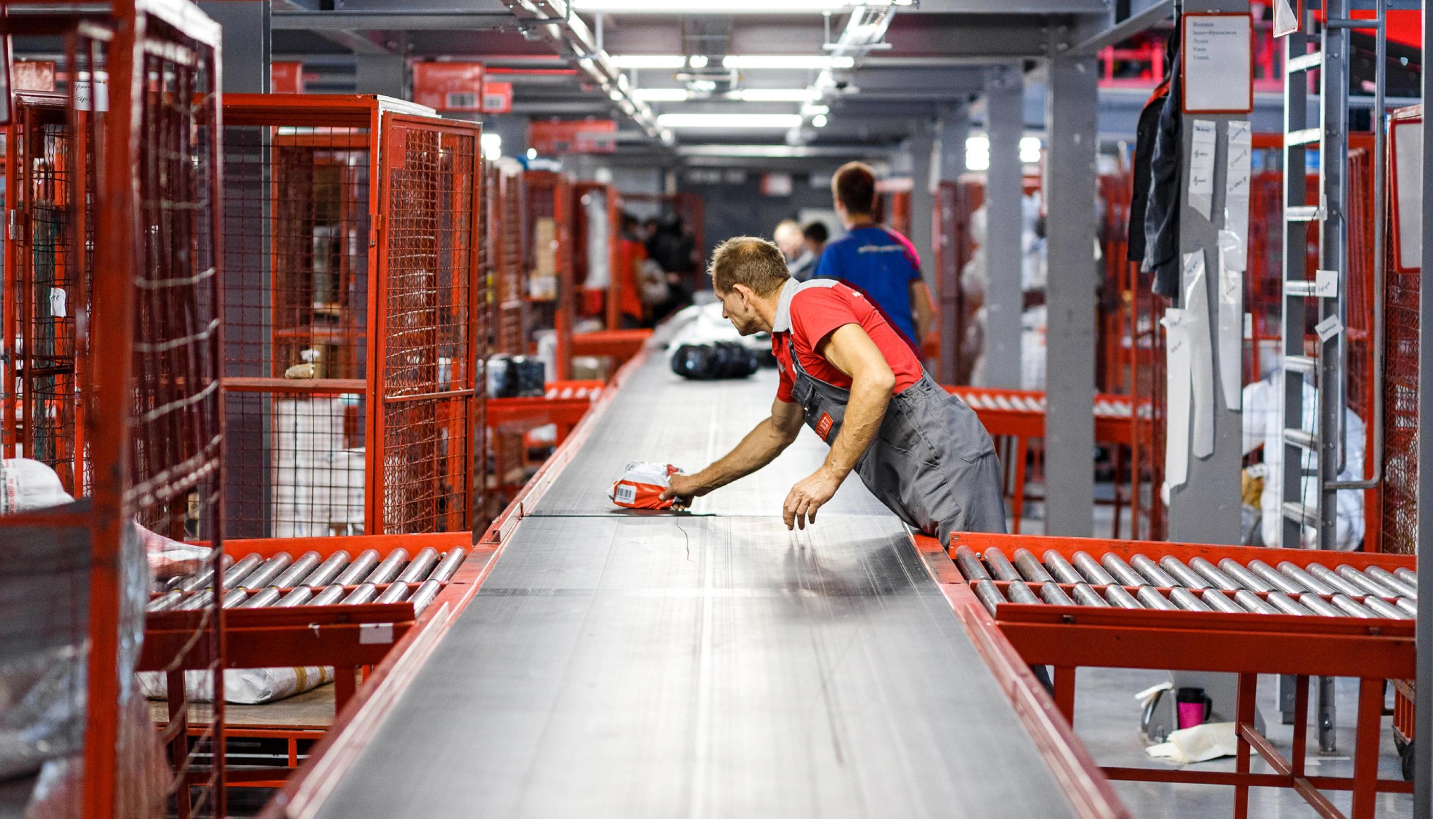 A high-resolution action photograph inside a Nova Post logistics terminal. A worker in grey overalls and a red branded shirt is captured in motion, placing a parcel onto a long, motorized conveyor belt system. The industrial setting features rows of red metal sorting cages and specialized rollers, highlighting the scale and human element behind the company's high-volume parcel processing.