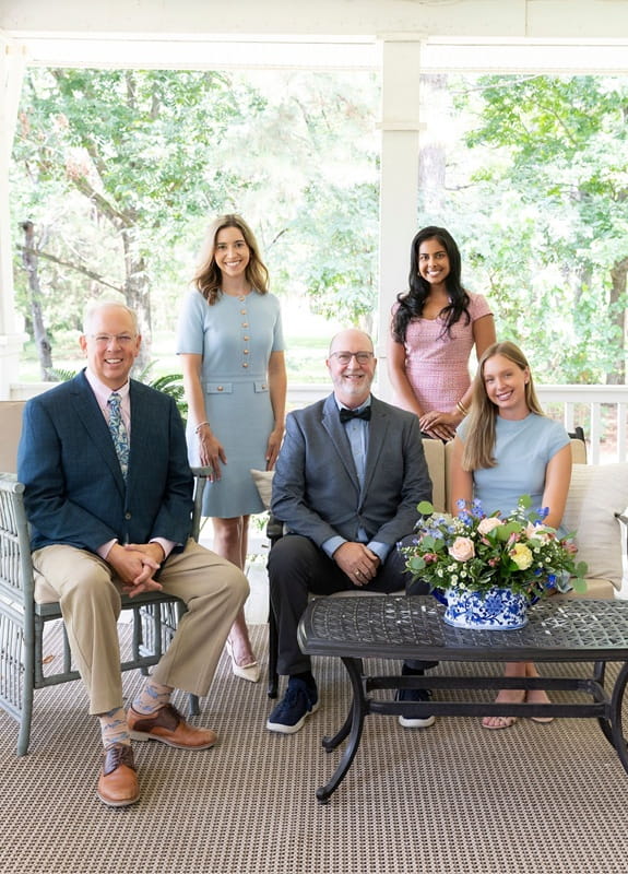 Group portrait of our dentists on a porch with trees in the background; two men sitting on chairs and three women standing or sitting around a black metal table with a floral centerpiece.