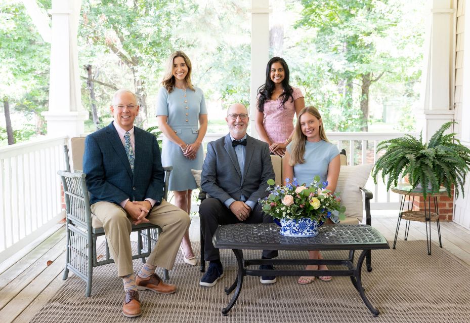 Five smiling people on a porch with greenery behind. Two men are seated on cushioned chairs, one in a blue blazer and tan pants and the other in a gray suit with a bow tie. Three women stand and sit between and behind them, wearing dresses in pastel colors. A black metal table with a blue and white vase of flowers is in front, and a potted fern is to the right.