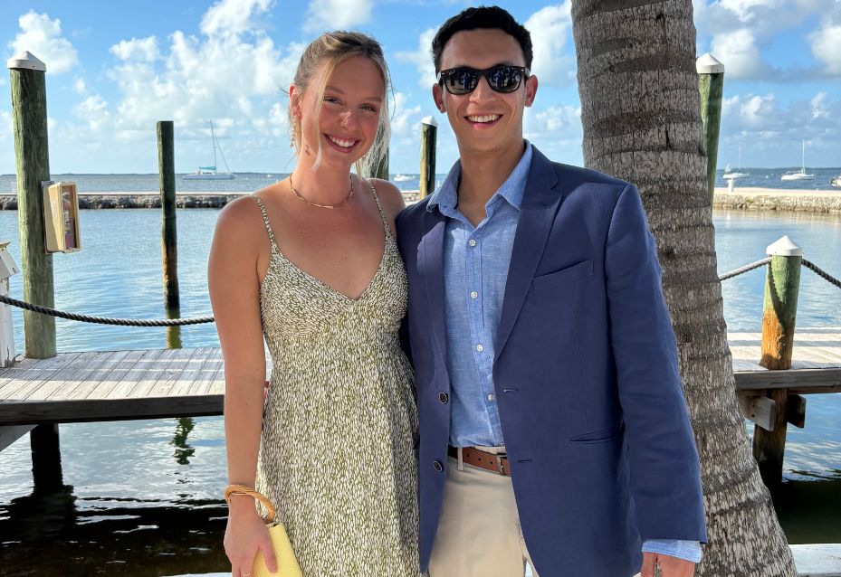 Two people stand side by side outdoors by the water, dressed in semi-formal summer clothing, smiling at the camera with a palm tree and dock in the background.