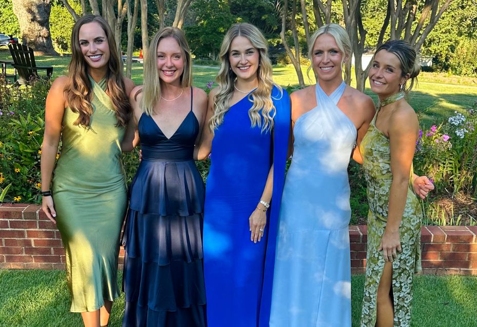 Five women in colorful formal dresses stand outdoors on grass in front of a flowerbed and trees, smiling at the camera.