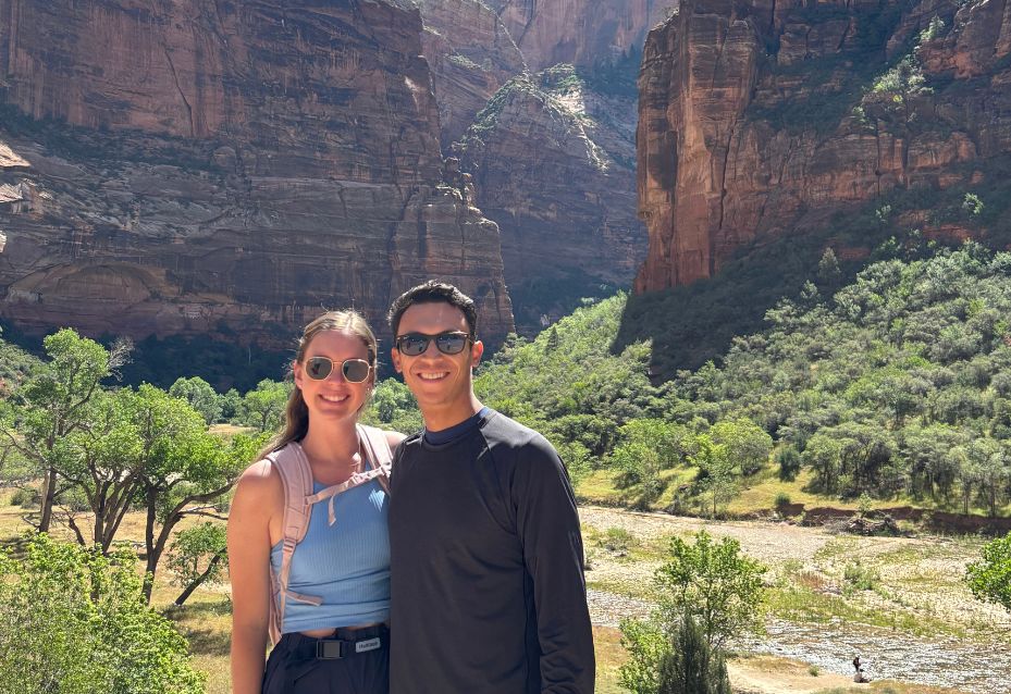 Two people stand side by side smiling in front of a sunlit canyon with tall rock walls, green trees, and a dry riverbed.