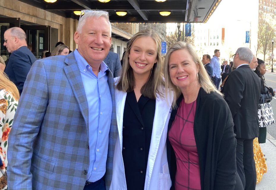 Three people stand closely together and smile at the camera outside a building, with other people and city street visible in the background.