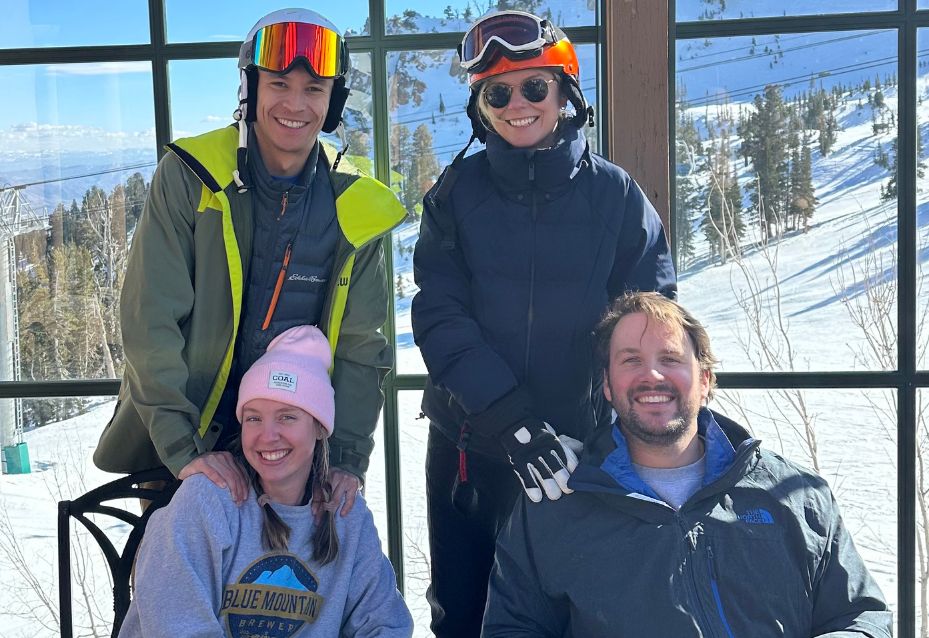 Four adults in winter clothing and ski helmets smiling together inside a ski lodge, with snow-covered ski slopes and pine trees visible through large windows in the background.