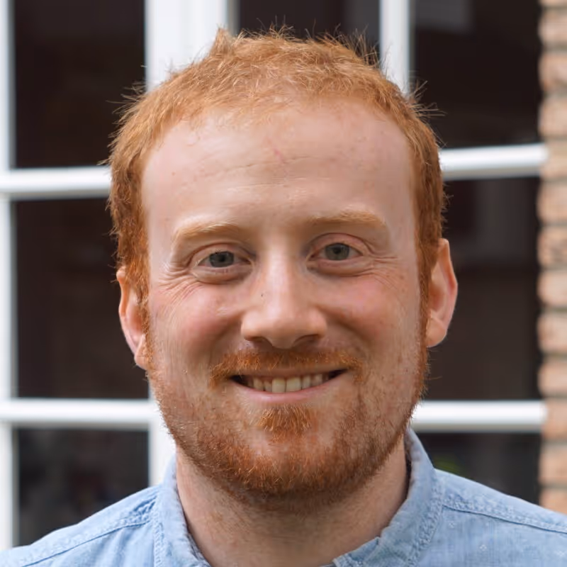 Smiling red-haired man with a short beard wearing a light blue shirt in front of a window and brick wall.