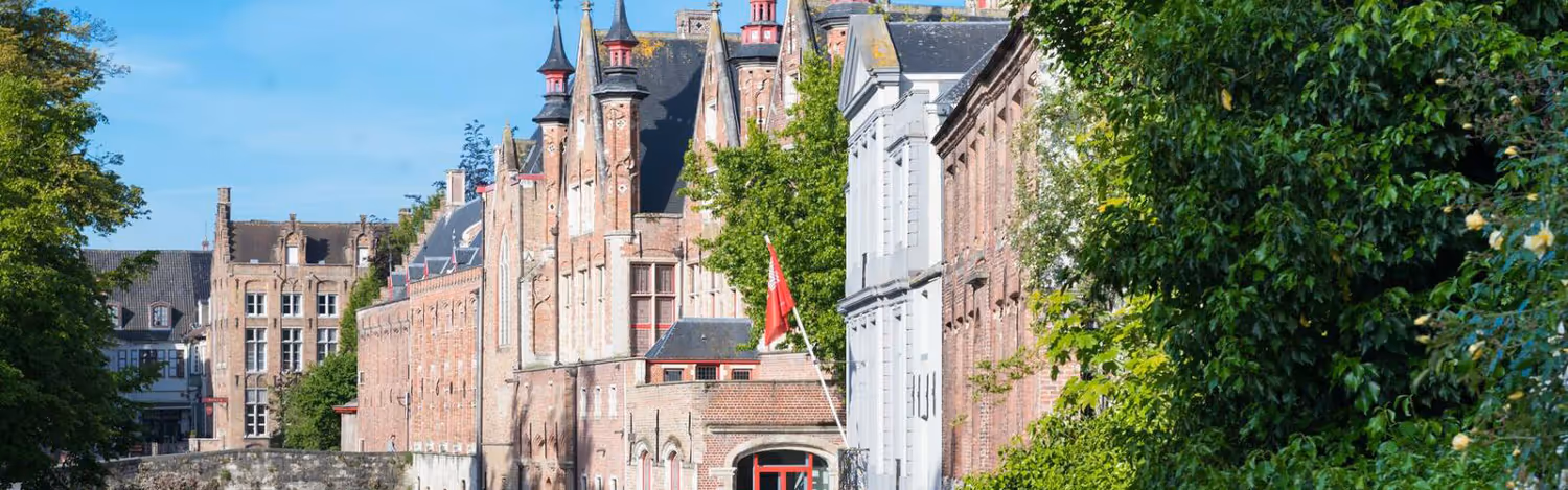 Historic red brick buildings with pointed rooftops along a canal with green trees and a clear blue sky.