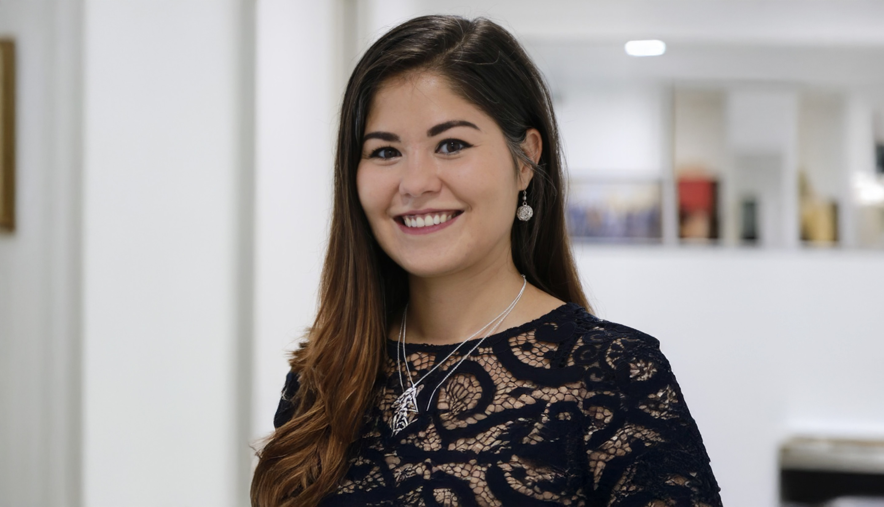 Smiling woman with long brown hair wearing a black lace top and silver jewelry in a bright indoor setting.
