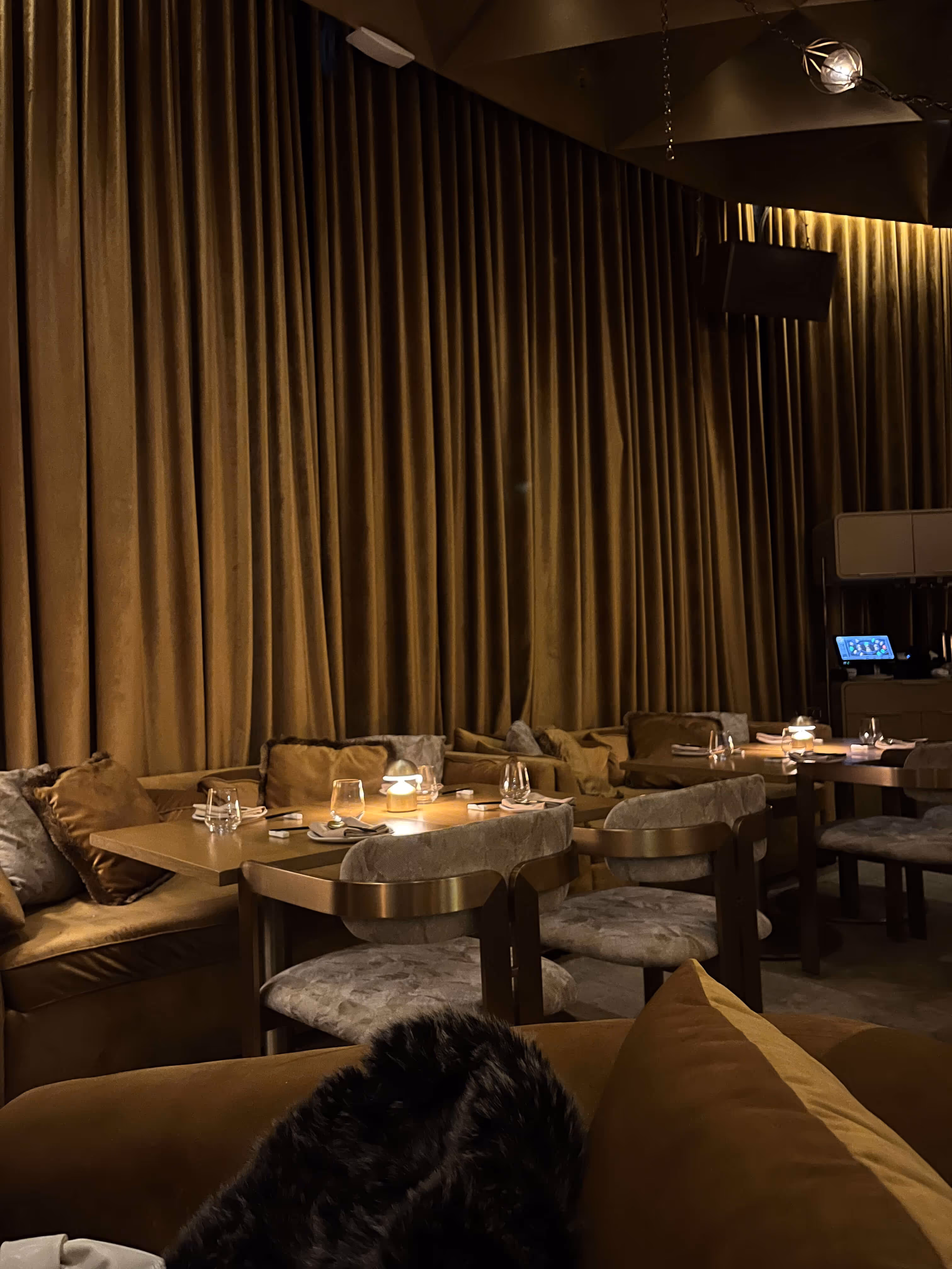 Dimly lit restaurant interior with beige velvet chairs, wooden tables, brown curtains, and soft glowing table lamps.