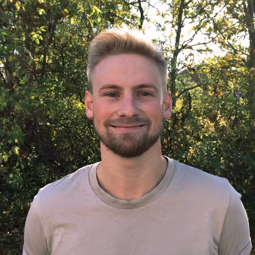 Smiling young man with short blond hair and beard standing outdoors in front of leafy trees.
