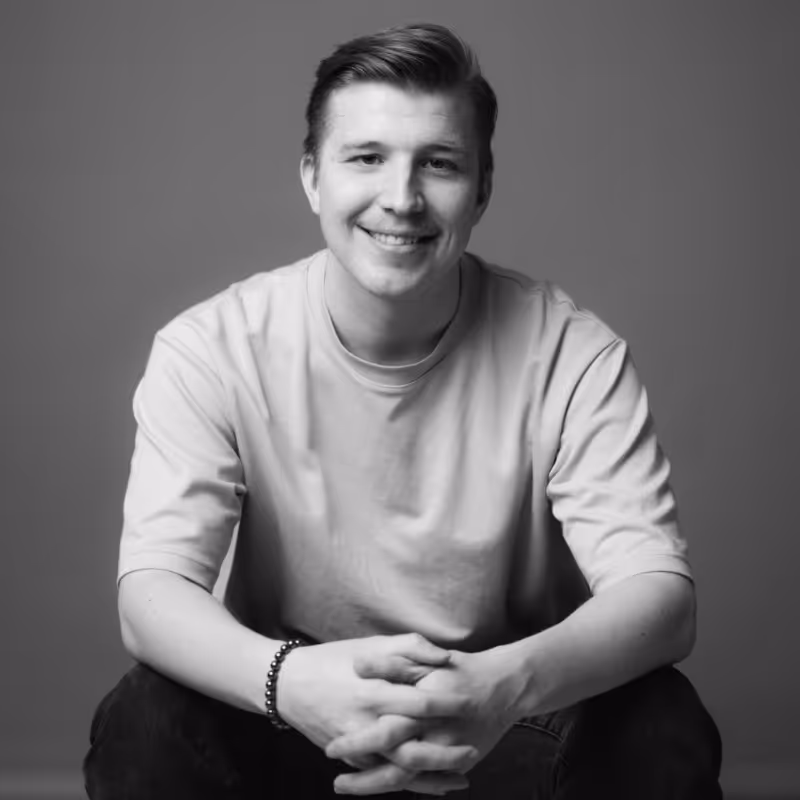 Young man sitting and smiling with hands clasped, wearing a light-colored t-shirt and bracelet.