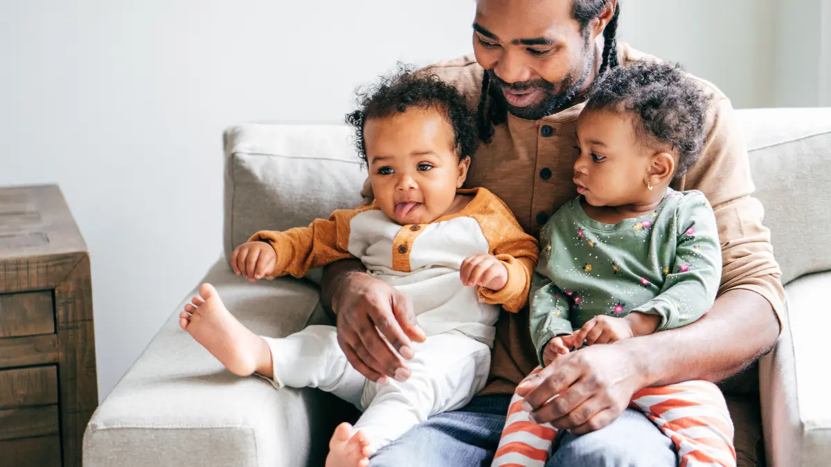 A father sits on a couch holding his two young children, representing the bonding time supported by FMLA and state paid family leave programs.