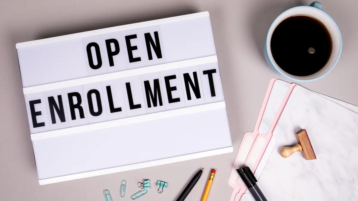Flat lay of office desk with coffee, pens, and a lightbox sign reading "Open Enrollment," representing open enrollment communication and planning.