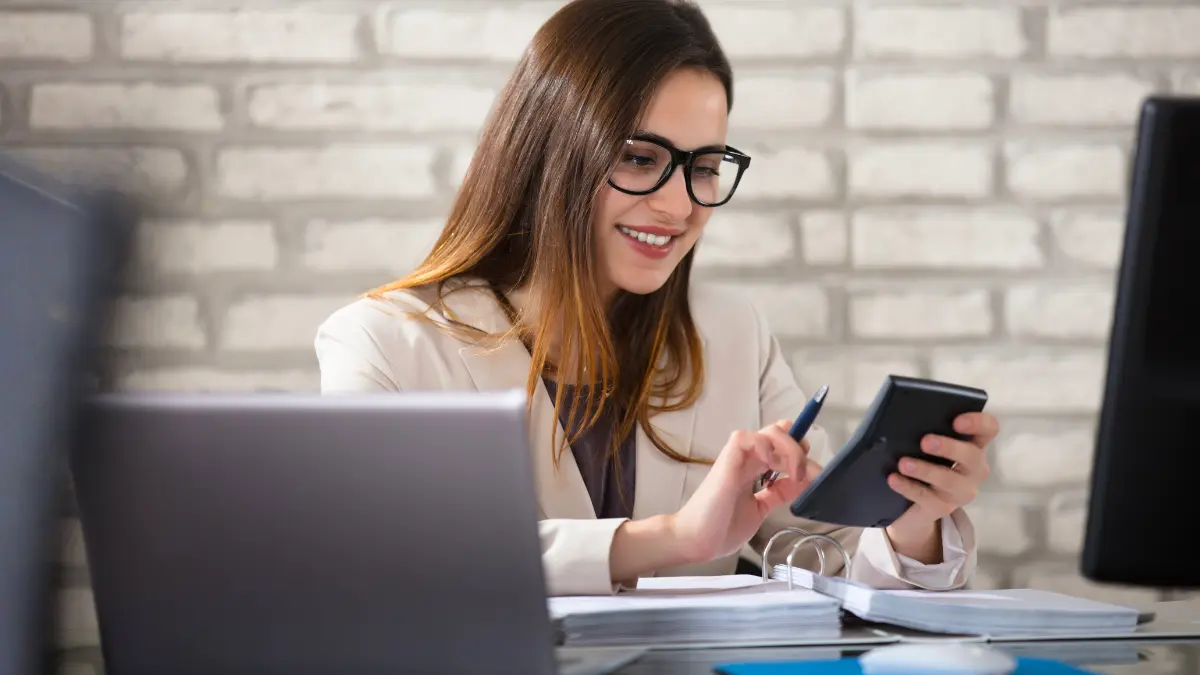A smiling woman wearing glasses sits at a desk reviewing financial documents, using a calculator and laptop in a bright office with a brick wall background.