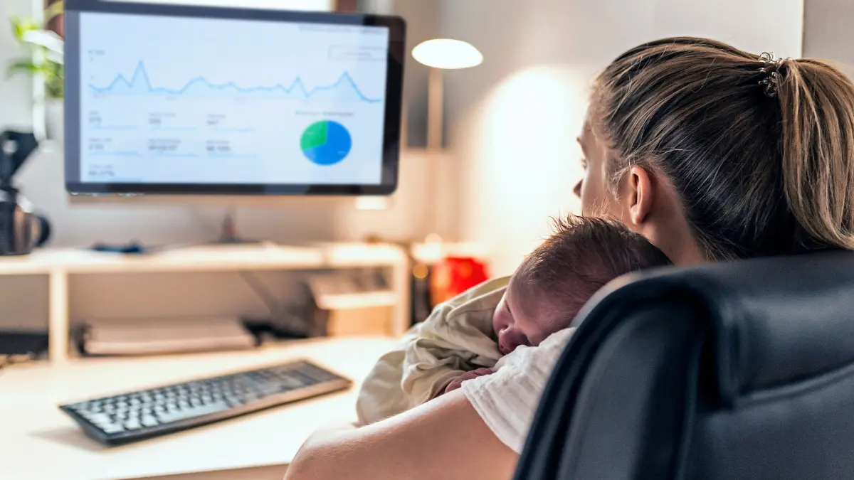 Woman working at a computer while holding a baby, illustrating the invisible labor and caregiving responsibilities many women balance alongside paid work.