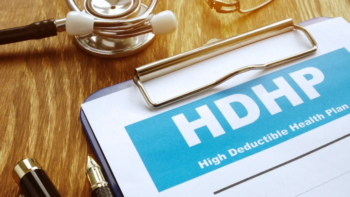 Close-up of a High Deductible Health Plan document on a clipboard with a stethoscope, symbolizing the decision employees face between an HDHP and traditional health insurance during open enrollment.