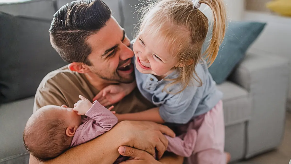 A father sits on the floor at home, cradling a newborn in one arm while his smiling toddler daughter leans in close, illustrating the value of parental leave for non-birthing parents and families.