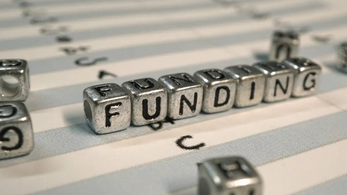 Metal letter cubes spelling "FUNDING" on a striped surface, representing employer health plan funding decisions