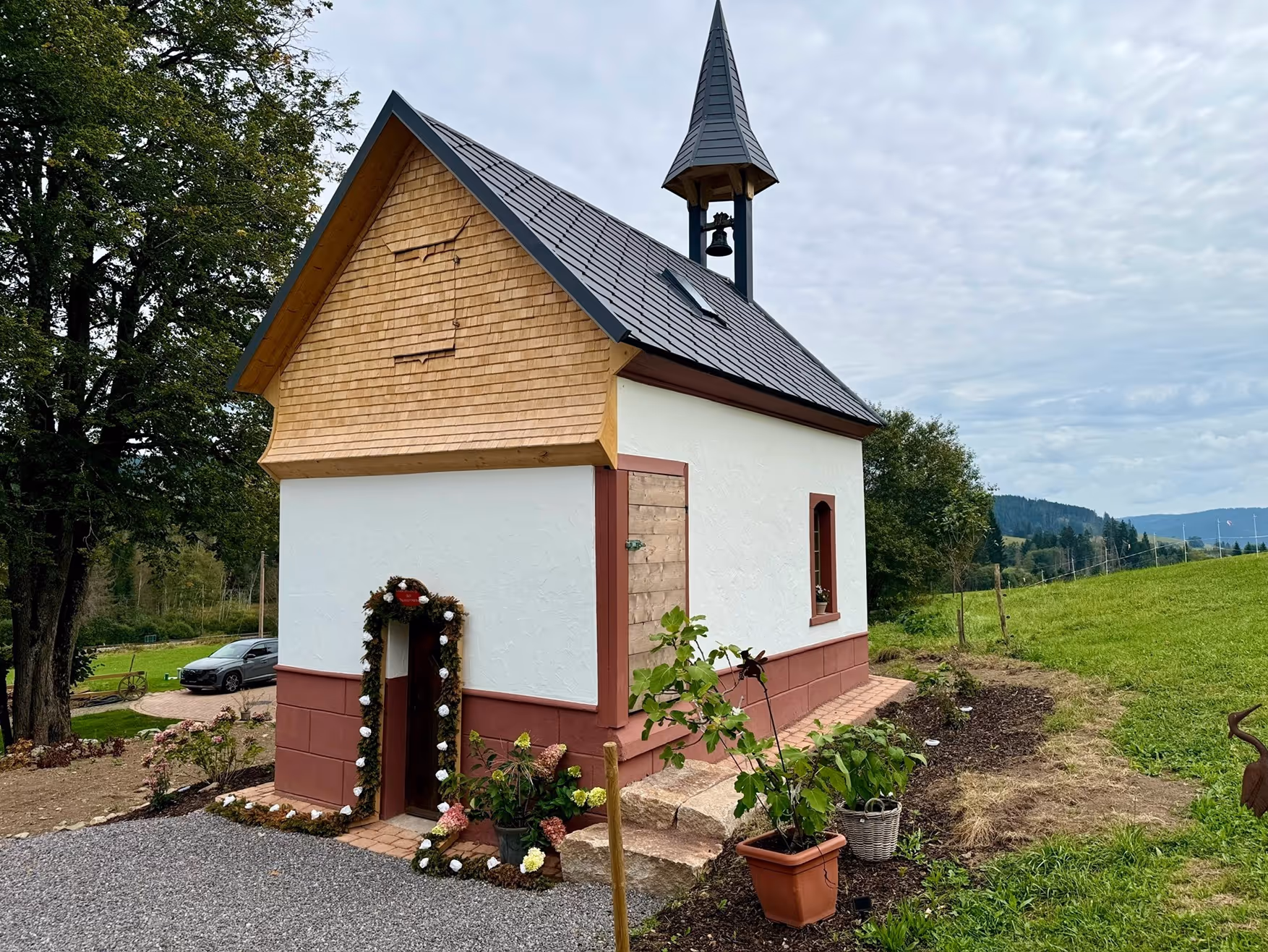 Frisch renovierte kleine Kapelle mit Holzschindeln und Glockenturm in ländlicher Umgebung, eingerahmt von Blumen und Bepflanzung.