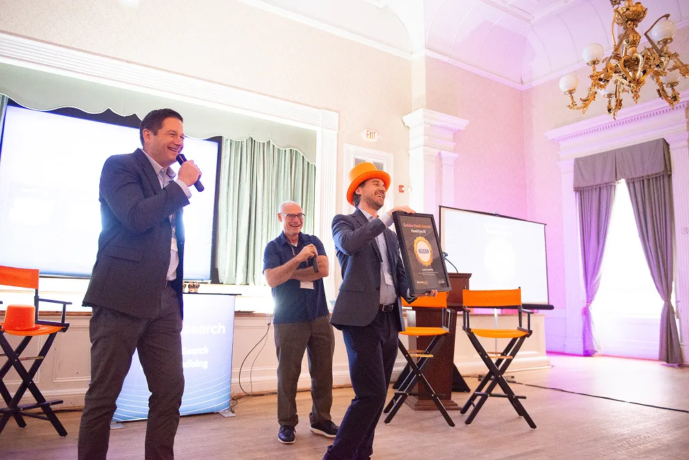 An award winner celebrates on stage wearing an orange hat while holding a Golden Hawk Award plaque during the HawkSearch Customer Summit 25.